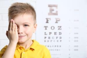 A little boy standing in front of a letter chart, covering one eye and smiling